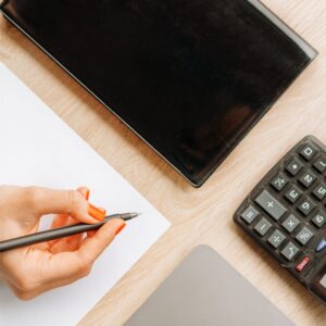 Mãos anotando informações em uma mesa de escritório com calculadora e documentos, em clima profissional e organizado