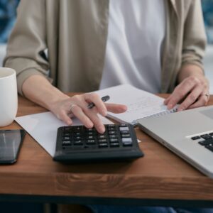 Pessoa usando calculadora e anotando em um caderno sobre mesa de madeira, em rotina financeira